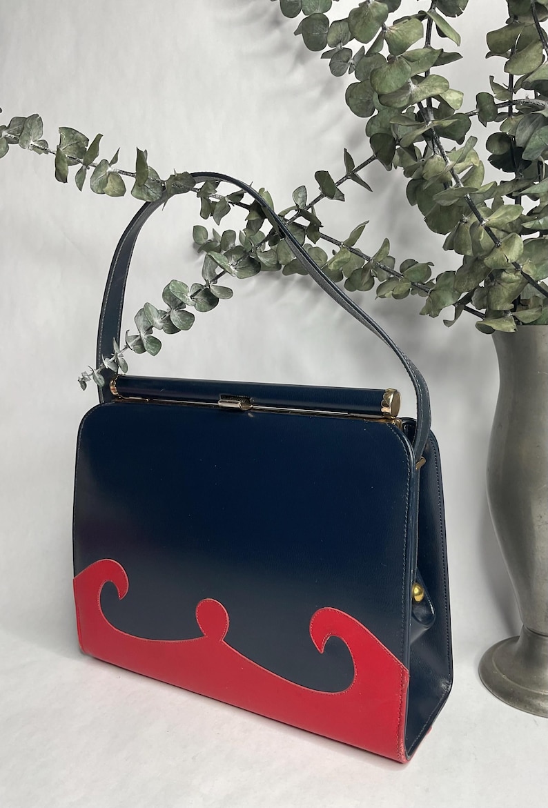 May include: A vintage navy blue handbag with a red wave design on the front. The bag has a rectangular shape, a top handle, and gold-colored hardware. The bag is displayed with a vase of greenery.