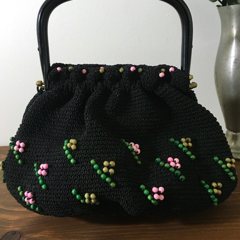 May include: A black crocheted handbag with a black plastic handle. The bag is decorated with pink and green beads in a floral pattern.