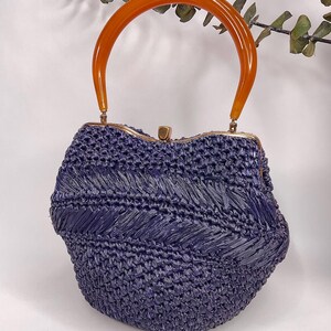 May include: A vintage-style handbag in a deep indigo hue, featuring a woven texture. The bag has a curved, amber-colored handle and a gold-toned clasp. The overall design evokes a retro aesthetic, suitable for carrying essentials.
