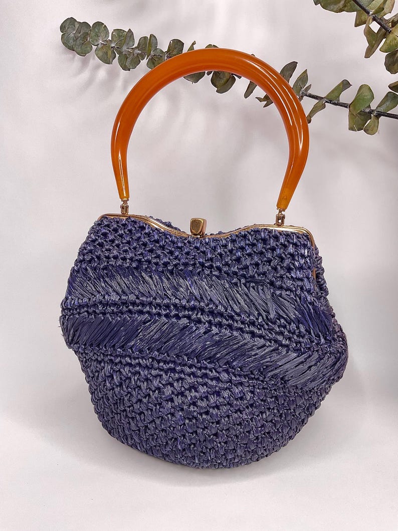May include: A vintage-style handbag in a deep indigo hue, featuring a woven texture. The bag has a curved, amber-colored handle and a gold-toned clasp. The overall design evokes a retro aesthetic, suitable for carrying essentials.