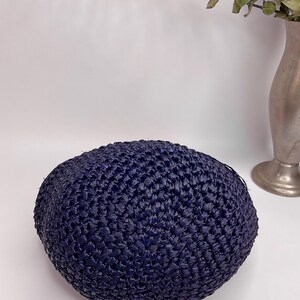 May include: A navy blue, woven, round hat. The hat is made of a textured material and has a slightly domed shape. The hat is sitting on a white surface.