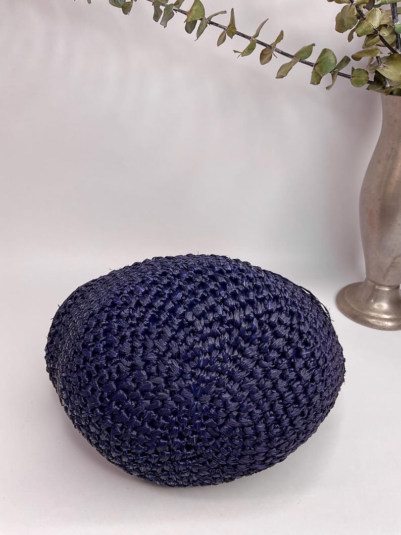 May include: A navy blue, woven, round hat. The hat is made of a textured material and has a slightly domed shape. The hat is sitting on a white surface.