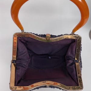 May include: Vintage navy blue handbag with a gold-tone frame and a large, curved orange handle. The interior is lined with dark purple fabric. The bag has a rectangular shape and a clasp closure. A classic accessory.