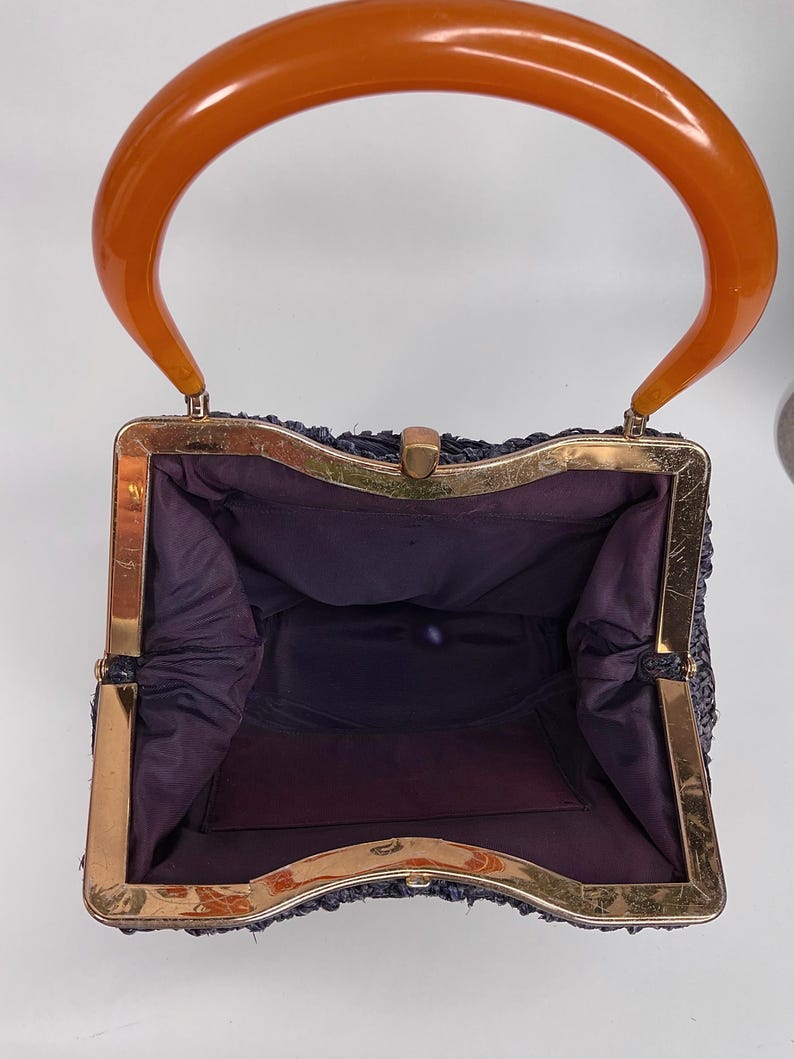 May include: Vintage navy blue handbag with a gold-tone frame and a large, curved orange handle. The interior is lined with dark purple fabric. The bag has a rectangular shape and a clasp closure. A classic accessory.