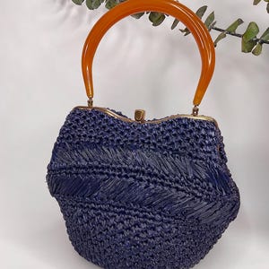 May include: A navy blue woven handbag with an orange, curved handle. The bag has a rounded shape with a gold-colored clasp. The woven texture creates a chevron pattern across the front of the bag. A stylish accessory.