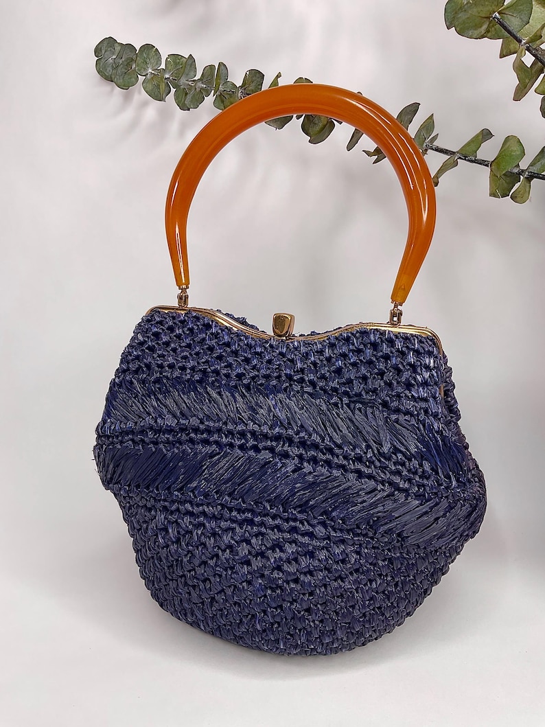 May include: A navy blue woven handbag with an orange, curved handle. The bag has a rounded shape with a gold-colored clasp. The woven texture creates a chevron pattern across the front of the bag. A stylish accessory.