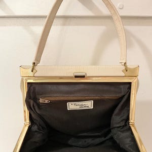以下が含まれることがあります： ゴールドフレームとハンドルが付いた、クリーム色のヴィンテージハンドバッグ。内側は暗く、「a Natali bag handbag」と書かれた小さなラベルが付いています。バッグは開いており、裏地とジッパーが見えます。