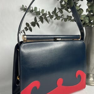 May include: A vintage navy blue handbag with a red wave design on the bottom. The bag has a gold clasp and handle attachments. The handle is navy blue. The bag is rectangular and has a structured shape. A classic, stylish accessory.