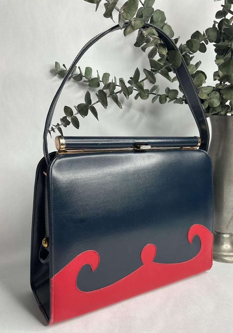 May include: A vintage navy blue handbag with a red wave design on the bottom. The bag has a gold clasp and handle attachments. The handle is navy blue. The bag is rectangular and has a structured shape. A classic, stylish accessory.