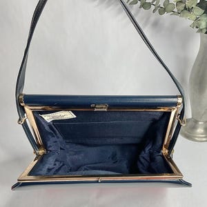 May include: Open navy blue handbag with a gold-colored frame and clasp. The interior is lined with navy blue fabric. The bag has a long, thin strap and a small tag inside. A vintage accessory.