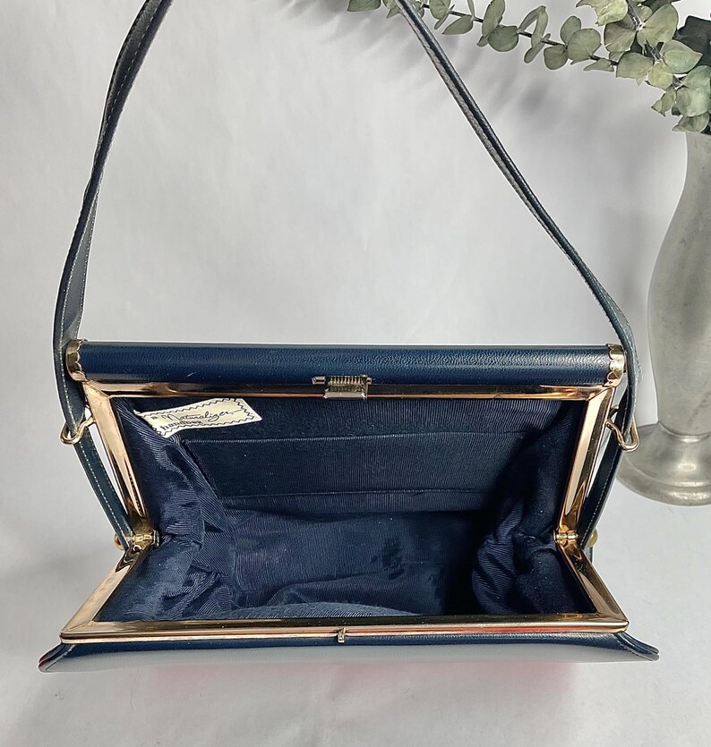 May include: Open navy blue handbag with a gold-colored frame and clasp. The interior is lined with navy blue fabric. The bag has a long, thin strap and a small tag inside. A vintage accessory.