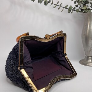 May include: Open vintage navy blue handbag with a gold-toned metal frame and clasp. The interior is lined with dark purple fabric. The handle is a light brown color. The bag has a woven texture.