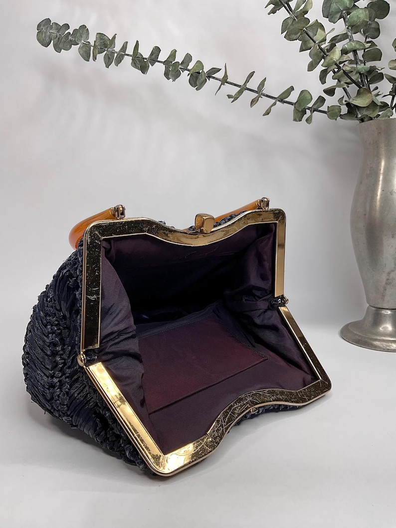 May include: Open vintage navy blue handbag with a gold-toned metal frame and clasp. The interior is lined with dark purple fabric. The handle is a light brown color. The bag has a woven texture.