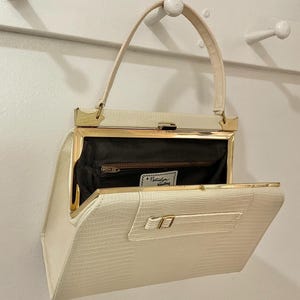 以下が含まれることがあります： クリーム色のヴィンテージハンドバッグ。表面はテクスチャード加工で、ゴールドトーンの金属アクセントが付いています。バッグはトップハンドル、長方形の形状で、開いたフラップからジッパーポケットとラベルが付いた黒い内側が見えます。