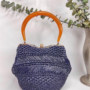 May include: A vintage navy blue woven handbag with an orange handle. The bag has a rounded shape with a gold-colored clasp. The handle is a curved, semi-circular shape. The bag is made of a woven material.
