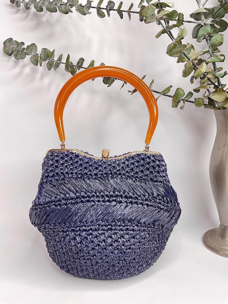 May include: A vintage navy blue woven handbag with an orange handle. The bag has a rounded shape with a gold-colored clasp. The handle is a curved, semi-circular shape. The bag is made of a woven material.