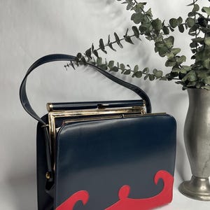 May include: A vintage navy blue handbag with a red wave design on the bottom. The bag has a gold-colored frame and a black handle. The bag is rectangular and appears to be made of leather or a similar material.