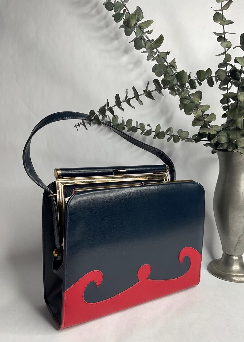 May include: A vintage navy blue handbag with a red wave design on the bottom. The bag has a gold-colored frame and a black handle. The bag is rectangular and appears to be made of leather or a similar material.