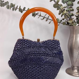 May include: A vintage navy blue woven handbag with an orange handle. The purse has a rounded shape and a gold-colored clasp. The handle is a curved, translucent orange color. The bag is set against a white background.
