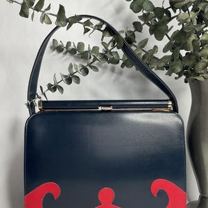May include: A vintage navy blue handbag with a red wave design on the bottom. The bag has a rectangular shape, a top handle, and a gold-colored clasp. The background includes greenery and a vase.