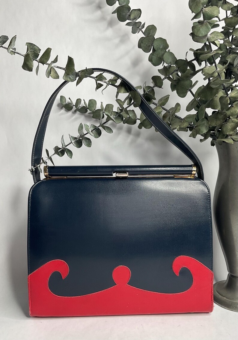 May include: A vintage navy blue handbag with a red wave design on the bottom. The bag has a rectangular shape, a top handle, and a gold-colored clasp. The background includes greenery and a vase.