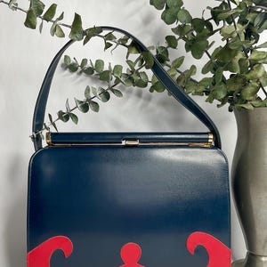 May include: A vintage navy blue handbag with a red decorative design on the bottom. The bag has a gold clasp and a navy blue handle. The bag is rectangular and appears to be made of leather.