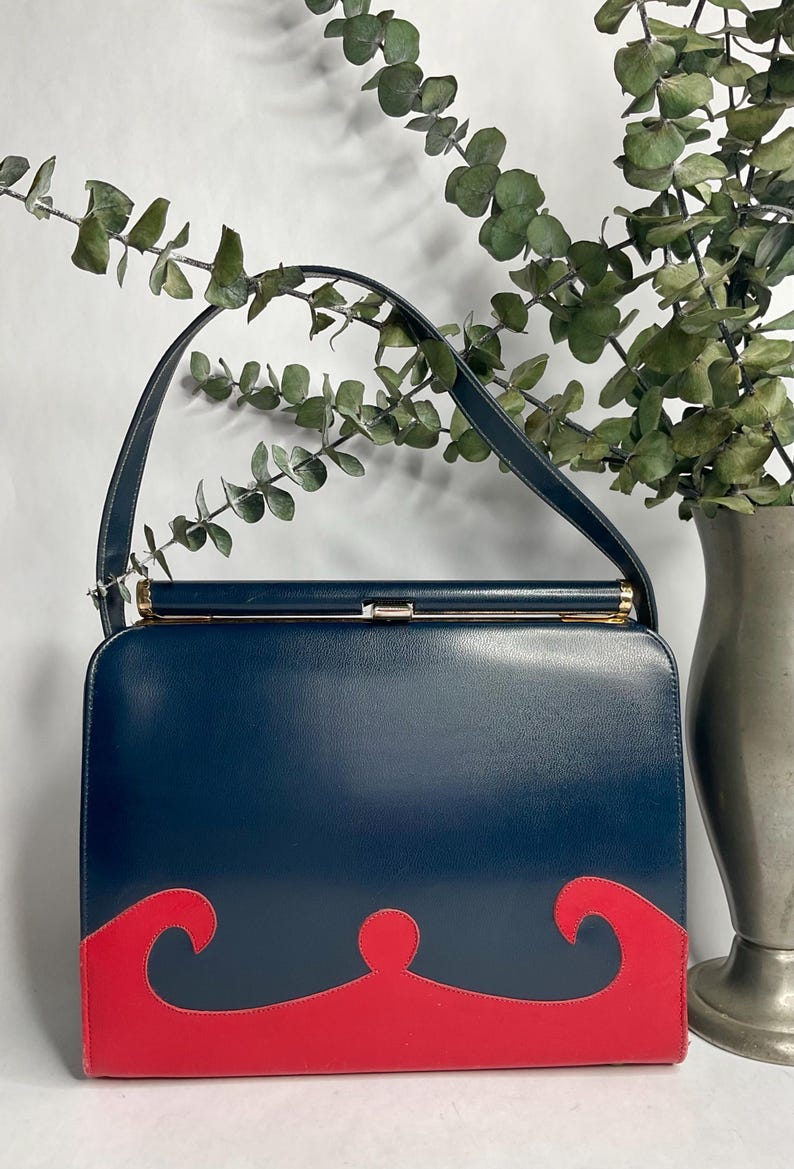 May include: A vintage navy blue handbag with a red decorative design on the bottom. The bag has a gold clasp and a navy blue handle. The bag is rectangular and appears to be made of leather.