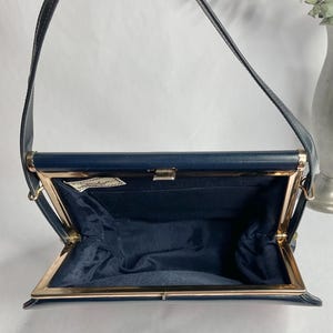 May include: Navy blue vintage handbag with a gold-toned clasp and frame. The bag has a structured shape with a long strap. The interior is lined with navy blue fabric. A small label is visible inside the bag.