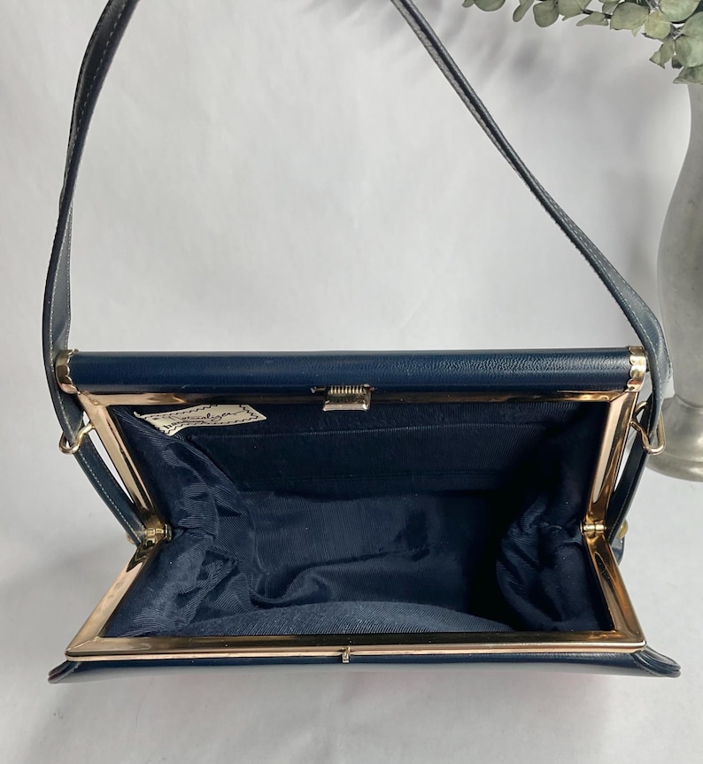 May include: Navy blue vintage handbag with a gold-toned clasp and frame. The bag has a structured shape with a long strap. The interior is lined with navy blue fabric. A small label is visible inside the bag.