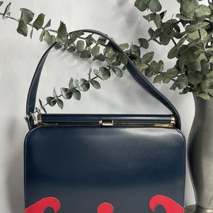 May include: A navy blue handbag with a red decorative design on the bottom. The bag has a gold clasp and a single handle. The background includes a vase with greenery.