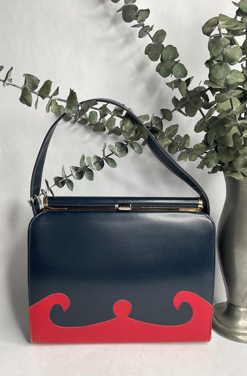May include: A navy blue handbag with a red decorative design on the bottom. The bag has a gold clasp and a single handle. The background includes a vase with greenery.