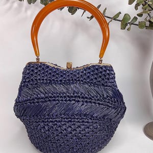 May include: A vintage navy blue woven handbag with a curved, amber-colored handle. The bag features a structured, rounded shape with a decorative woven pattern. The handle is attached with gold-toned hardware, creating a stylish accessory.