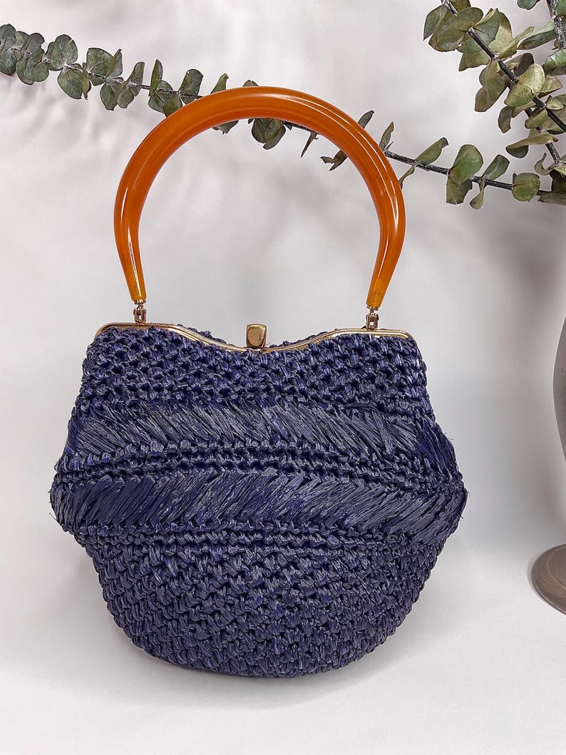 May include: A vintage navy blue woven handbag with a curved, amber-colored handle. The bag features a structured, rounded shape with a decorative woven pattern. The handle is attached with gold-toned hardware, creating a stylish accessory.