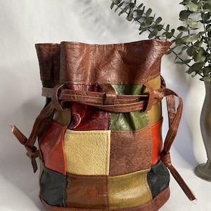 May include: A patchwork leather bucket bag in shades of brown, red, green, and cream. The bag features a drawstring closure and a strap that can be tied. The bag is a unique accessory.