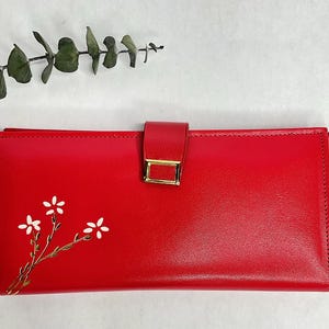 May include: A bright red wallet with a gold-colored clasp. The wallet features a floral design with white flowers and gold stems. The wallet is rectangular and has a flap closure.