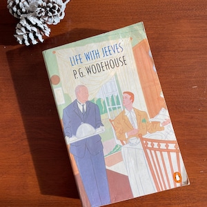 May include: A vintage paperback book titled "Life with Jeeves" by P.G. Wodehouse. The cover features an illustration of two men, one in a suit holding a tray. The book has a Penguin logo and is set on a wooden surface with decorative pinecones.