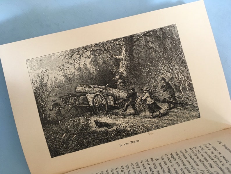 May include: An open book displays a black and white illustration titled "In the Woods." The image depicts a horse-drawn cart loaded with logs, being pulled by people through a wooded area. A dog runs nearby.