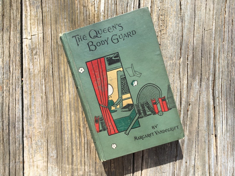 May include: Vintage book titled "The Queen's Body Guard" by Margaret Vandegrift. The book cover is a muted green with illustrations of a tennis racket, books, and a window with a red curtain. The book is resting on a weathered wooden surface.