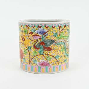 Op de afbeelding: Een gele keramische cilindervaas met een kleurrijk bloemen- en vogelontwerp. De vaas heeft een roze, groene en gele rand.