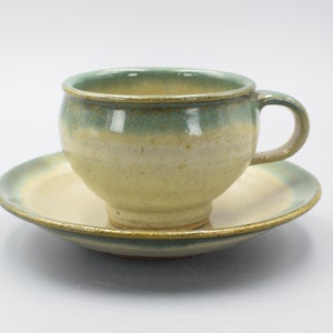 Pode incluir: Um conjunto de chávena e pires de cerâmica com um esmalte verde claro e amarelo. A chávena tem uma forma arredondada e uma pega. O pires é uma forma redonda simples.