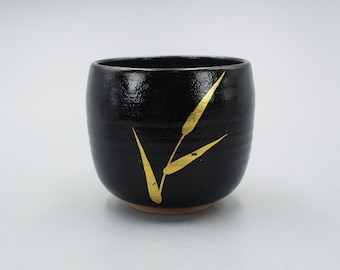 May include: A black ceramic teacup with a gold leaf design. The cup is round and has a slightly flared lip.