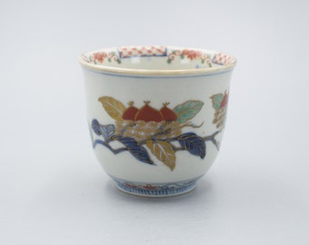 May include: A white porcelain teacup with a floral design. The cup is decorated with red, blue, and gold paint. The design features a branch with leaves and flowers.