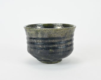 May include: A dark green ceramic teacup with a textured, slightly uneven surface. The cup has a wide, rounded base and a slightly flared rim.