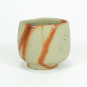 Puede incluir: Una taza de cerámica hecha a mano con un exterior verde claro moteado. Dos bandas diagonales de esmalte marrón rojizo adornan la taza. La taza tiene un cuerpo redondeado y una pequeña base con pie, ideal para té o café.