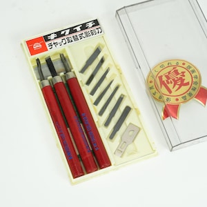 May include: A set of wood carving tools with red handles and black blades, packaged in a cream-colored case with extra blades. The case is next to a clear plastic box with a gold and red award seal.