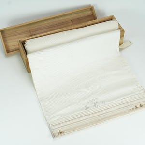 Può includere: Una scatola di legno con coperchio, contenente un tessuto bianco arrotolato con un sottile motivo a onde. Il tessuto è parzialmente srotolato, rivelando un design dettagliato e testo giapponese. La scatola è in legno chiaro.