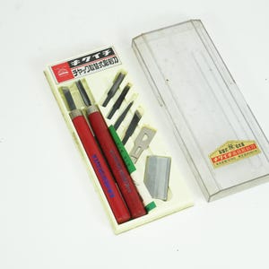 May include: A set of wood carving tools in a cream-coloured case with a clear plastic lid. The set includes two red-handled chisels, various blades, and the text "キイチ" in Japanese. The tools are arranged on a cream-coloured base.