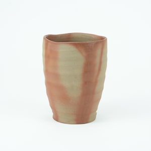 Puede incluir: Un vaso de cerámica hecho a mano con una forma orgánica única. La taza de color beige presenta patrones arremolinados de color marrón rojizo. El borde tiene un borde ligeramente irregular. Esta taza de gres es adecuada para bebidas.