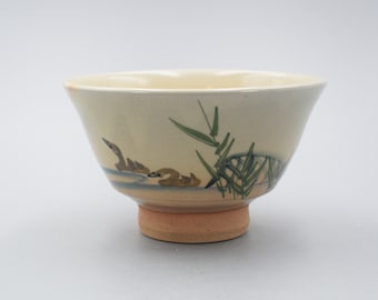 May include: A small, cream-colored ceramic bowl with a brown and green design of birds and reeds. The bowl has a rounded bottom and a slightly flared rim.