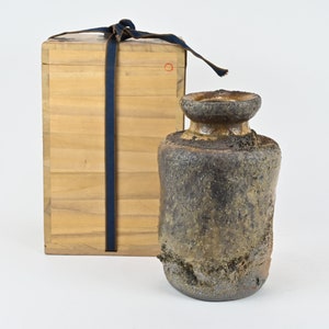 May include: A dark brown ceramic vase with a rough, textured surface. The vase has a wide mouth and a narrow neck. It is sitting on a white surface next to a wooden box with a blue ribbon.